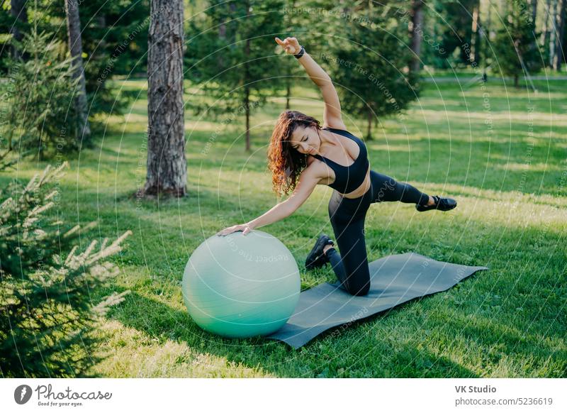 Flexible aktive Frau in Sportkleidung macht Fitness-Übungen auf karemat mit fitball, hebt die Arme und atmet frische Luft im Wald hat Morgen Workout Posen über Natur Hintergrund. Gesunder Lebensstil