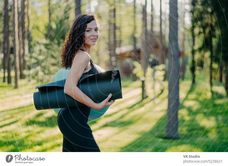 Sideways Schuss von sportlichen schlanke junge Frau in schwarzen Leggings gekleidet, trägt aufgerollt karemat unter dem Arm, posiert gegen grünes Gras und Bäume Hintergrund, gehen, um Übungen mit Fitness-Ball haben
