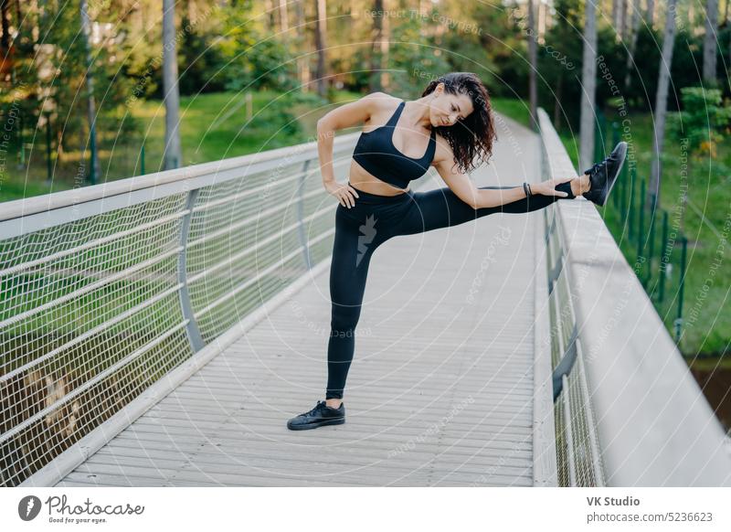 Entschlossene Sportlerin streckt die Beine auf einer Brücke, trägt aktive Kleidung, zeigt gute Flexibilität, posiert im Freien vor einem Waldhintergrund. Frau Läufer bereitet sich auf Joggen, hat Sommer-Workout