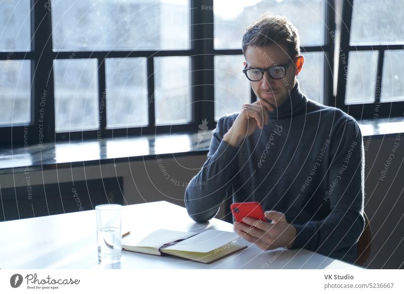 Junger deutscher Geschäftsmann, der am Schreibtisch sitzend Internetnachrichten liest oder E-Mails auf seinem Smartphone abruft jung Deutsch Mann Mobile Telefon