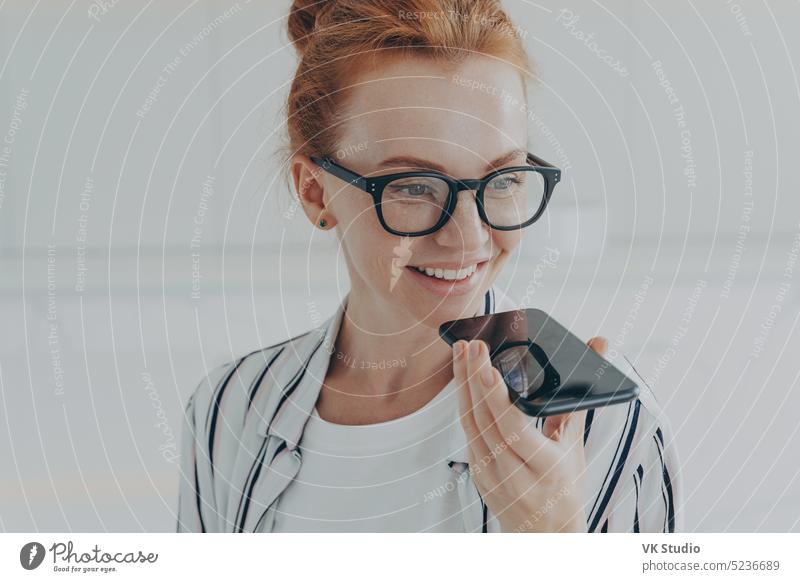 Rothaarige Frau mit durchsichtiger Brille spricht mit virtuellem Assistenten, der das Mobiltelefon an den Mund hält Person Gesicht Technik & Technologie Stimme