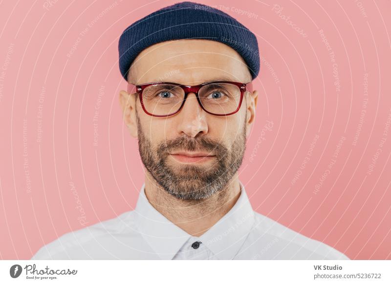 Nahaufnahme eines selbstbewussten männlichen Angestellten mit dichtem Bart und Schnurrbart, der eine durchsichtige Brille, einen Hut und ein weißes Hemd trägt, vor einem rosa Hintergrund steht und aufmerksam in die Kamera blickt. Gesichtsausdruck