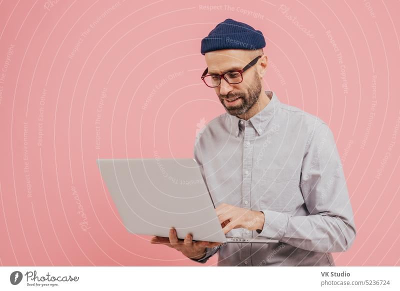 Professionelle männliche Arbeiter sucht nach interessanten Film zu sehen, verwendet Gadget, Typen Informationen auf Laptop-Computer, trägt formale Kleidung, isoliert über rosa Hintergrund. Online-Kommunikation