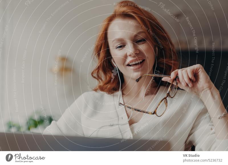 Selbstbewusste, zufriedene, rothaarige Frau, die sich auf einen Laptop konzentriert, sieht sich ein Video oder einen Film auf einem Laptop an, genießt einen freundlichen Videoanruf, hält eine durchsichtige Brille, nutzt eine kostenlose Internetverbindung