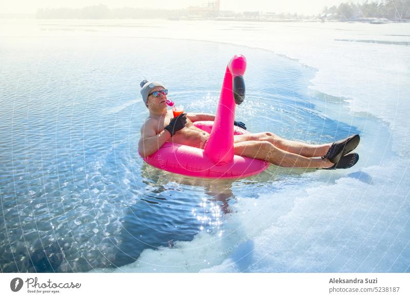 Ein Mann, der im Winter in Finnland in einem Eisloch schwimmt, schwebt auf einem rosa aufblasbaren Flamingo mit einem Cocktail in der Hand. Urlaubsoptionen, die vom Sommer träumen.