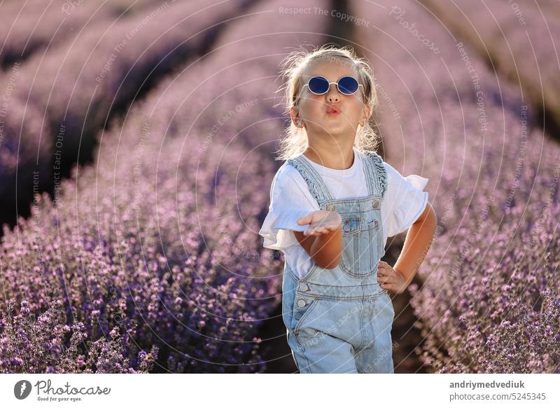Adorable glückliches Kind Mädchen hat Spaß und bläst Luft Kuss auf Kamera in Lavendelfeld auf Sommer warmen Tag. Hyperaktiv kleines Kind in Sonnenbrille auf die Natur. Internationaler Tag der Kinder, Valentinstag