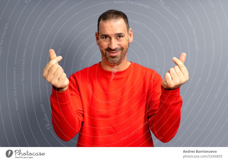 Bärtiger hispanischer Mann in den 40ern, der einen roten Pullover trägt und die Geste des Geldes macht, indem er seine Finger mit einem gierigen Gesicht reibt, isoliert auf grauem Hintergrund.