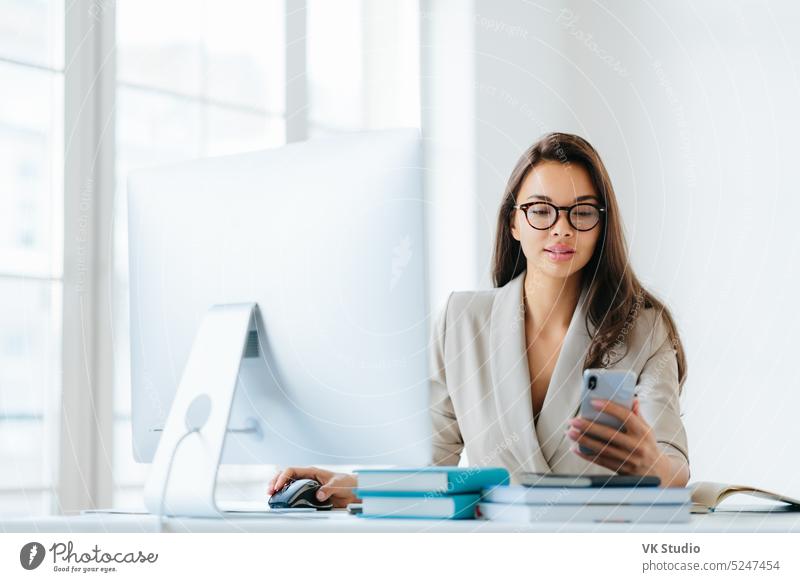 Elegante Büroangestellte benutzt Handy und Computer gleichzeitig, sendet Nachrichten und chattet mit Kunden online, verfolgt Nachrichten in Netzwerken, sitzt am Schreibtisch mit einem Stapel Notizblöcke und Bücher