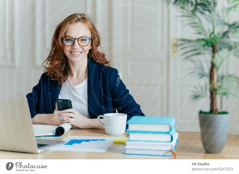 Foto einer rothaarigen, lächelnden jungen Frau, die eine App auf ihrem Mobiltelefon installiert, den Newsfeed prüft, aus der Ferne an einem Laptop arbeitet und im Coworking Space mit einer Tasse Getränk, Büchern und Papierdokumenten posiert