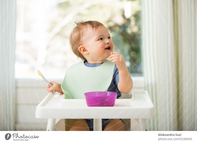 Süßes Baby isst erste feste Nahrung, Kleinkind sitzt im Hochstuhl. Kind probiert Gemüse am Tisch, entdeckt neue Lebensmittel. Gemütliche Küche Interieur. Gesundes Essen Konzept.