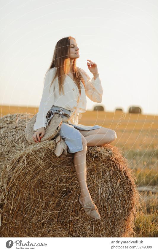 Junge Frau mit langen Haaren, in Jeansrock, hellem Hemd und Strohsack in der Hand, sitzt auf einem Ballen auf einem Feld im Sommer. Weibliches Porträt in natürlicher ländlicher Szene. Ökotourismus-Konzept.