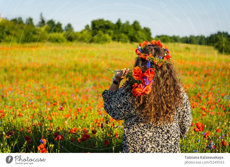 Langhaarige lockige Frau in Übergröße steht inmitten einer Mohnwiese mit einem Kranz aus Mohn- und Kornblumen und hält einen Strauß Mohnblumen. langhaarig