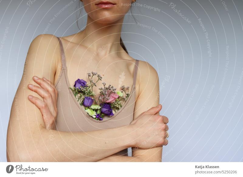 Eine Junge Frau mit gekreuzten Armen und in einem beigen Brusthalter in dem bunte Blumen stecken. gekreuzte Arme Blumen im Brusthalter beider Brusthalter Bra