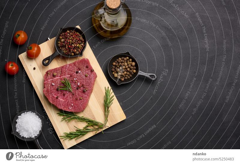 Rohes Stück Rindfleisch mit Gewürzen Pfeffer, Rosmarinzweig, Salz und Olivenöl auf einem Holzbrett, schwarzer Hintergrund. Platz zum Kopieren roh Paprika