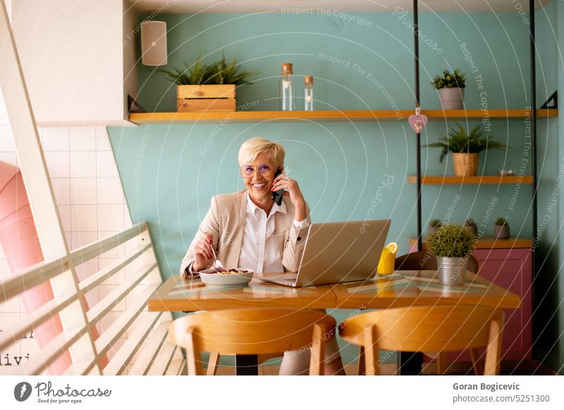 Ältere Frau, die ein Mobiltelefon benutzt, während sie am Laptop arbeitet und ein gesundes Frühstück im Café einnimmt gealtert Anruf Kaffee Kaffeehaus