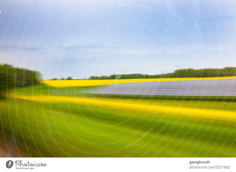 solare Rapsfahrt | Foto aus einer Bahn- oder Autofahrt vorbei an einer Wiese und Rapsfeld, sowie Solarmodulen. Im Hintergrund sind Windkraftwerke erkennbar, im Vordergrund Farbspektren des Verbundglases.