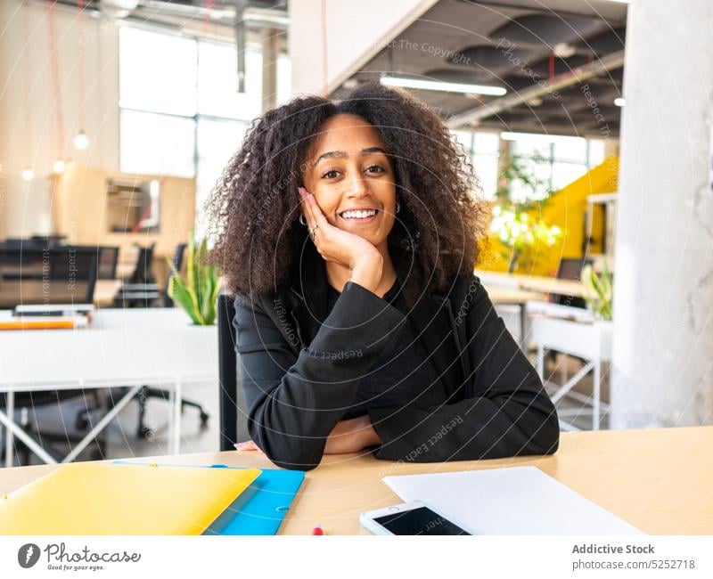 Verträumte schwarze Mitarbeiterin sitzt im Büro Frau sich auf die Hand lehnen verträumt Tisch Smartphone Unternehmen Arbeitsbereich Beruf nachdenken Lächeln