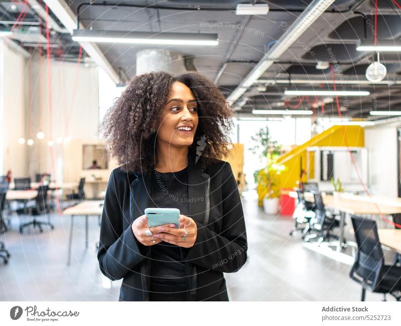 Positive schwarze Frau benutzt Smartphone in modernem Büro benutzend Nachricht Talkrunde Arbeitsbereich Mitarbeiter Unternehmen Gerät positiv Lächeln jung