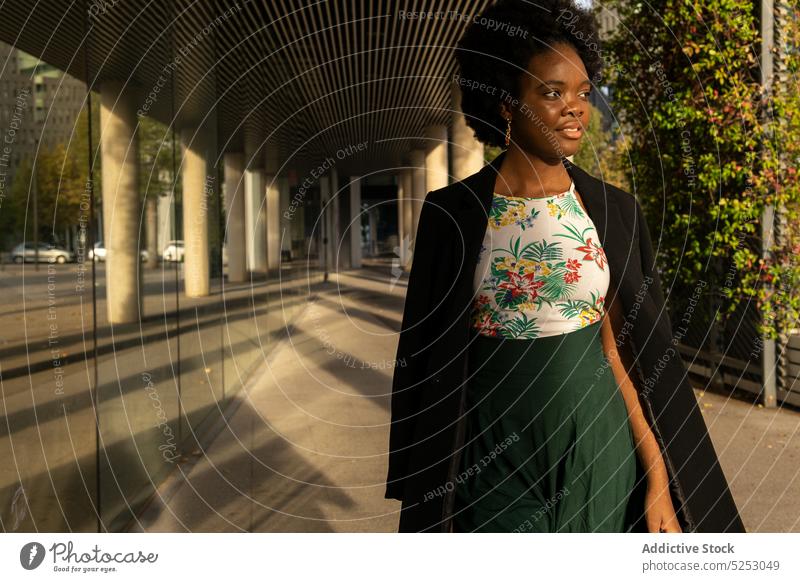 Stilvolle ethnische Frau in der Nähe eines Gebäudes stehend besinnlich verträumt nachdenklich urban Großstadt Sonne Afroamerikaner Straße Windstille jung