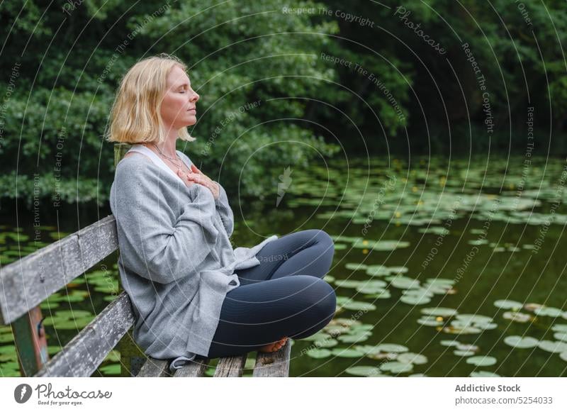 Ruhige Frau sitzt in Lotus-Pose in der Nähe von Teich padmasana meditieren Bank Lotosblatt Augen geschlossen Erholung Lebensmitte genießen Sprit See Yoga Asana