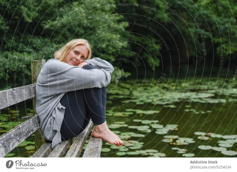 Ruhige Frau umarmt Knie sitzen auf Bank in der Nähe von Teich Knie umarmend ruhen Lotosblatt Baum Park Erholung Landschaft nachdenken Lebensmitte genießen Kälte
