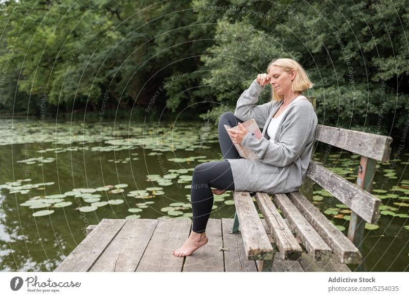 Nachdenkliche Frau mit Notebook auf einer Bank am See sitzend lesen Hinweis Information Buch Schönschreibheft Park Natur Baum nachdenklich Denken Gedanke