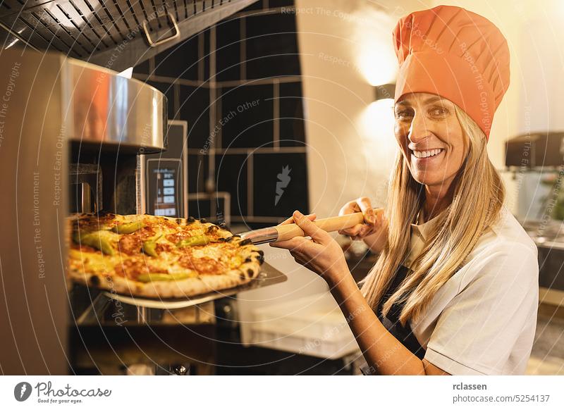 traditionelle Pizzabäckerin, die eine frische Pizza aus dem Steinofen auf einer Schale in einer italienischen Pizzeria hält Frau Hut Holzfeuer Ziegelofen