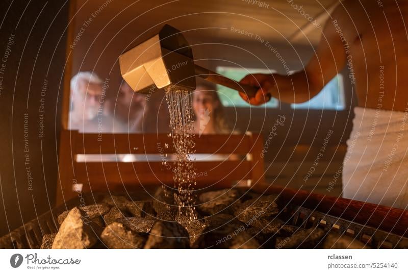 Mann gießt Wasser auf heiße Steine mit regen Löffel in Sauna Zimmer mit einer Gruppe von Menschen. Dampf und Wasser auf den Steinen, Spa- und Wellness-Konzept, Entspannung in der heißen finnischen Sauna.