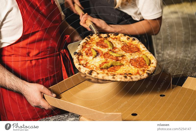 Pizzabäcker, die eine frische Salami-Peperoni-Pizza aus dem Steinofen in einen Karton für die Auslieferung in einer italienischen Pizzeria ziehen Hütte
