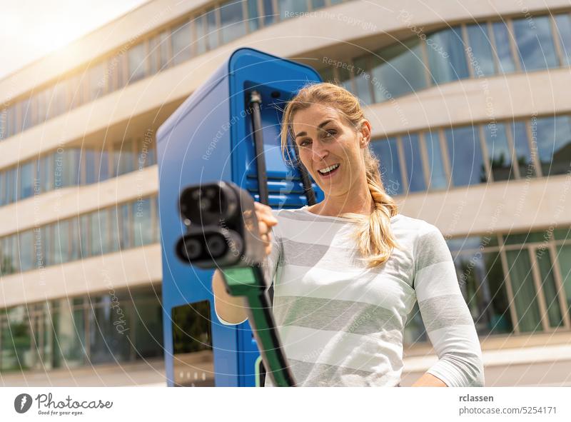 Eine Frau hält einen DC CCS2 EV-Ladestecker an einem Hypercharger oder Supercharger, um ihr Auto aufzuladen. Glück nfc Debitkarte Zahlung bezahlen Postkarte