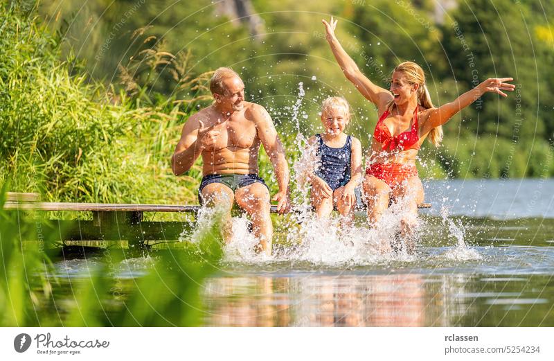 Glückliche Familie badet und spritzt Wasser mit dem Fuß am Steg auf einem Fluss im Sommer Schilfrohr Urlaub Badebekleidung Bikini Bayern Deutschland