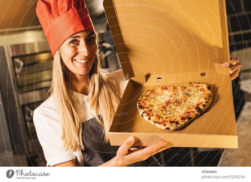 Pizzabäckerin hält eine frische Käsepizza in Herzform aus dem Steinofen in einer Schachtel für die Lieferung in einer italienischen Pizzeria Lächeln