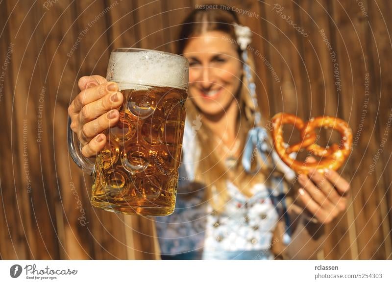 Fröhliche Frau stößt mit einem großen Krug Bier in traditioneller bayerischer Tracht an, in der anderen Hand hält sie eine Brezel im bayerischen Biergarten oder auf dem Oktoberfest
