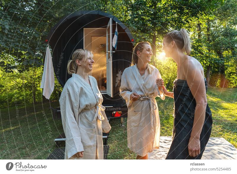 Drei Freundinnen unterhalten sich und stehen vor einer Holzfass-Sauna im Sommer in Norwegen. Sie sind entspannt, lachen und genießen den Urlaub, während sie in der finnischen Saunakabine entspannen.
