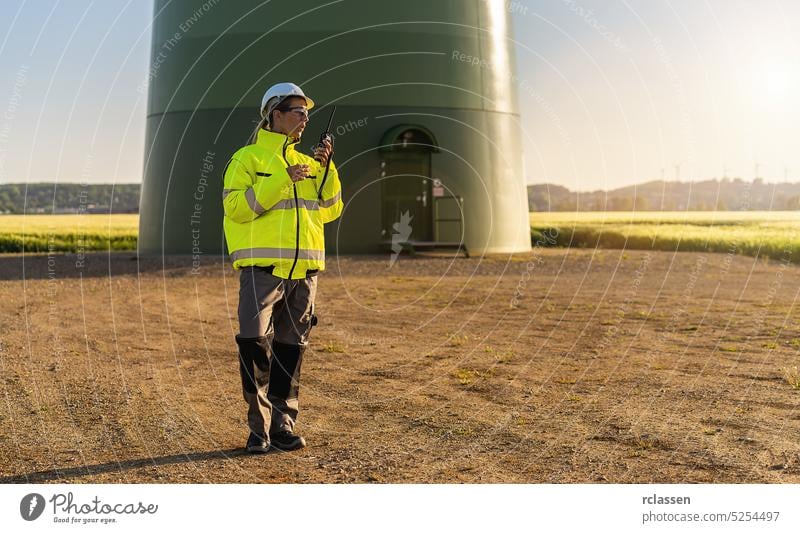 Ingenieurin mit Sicherheitsjacke vor einer Windturbine, die ein Walkie-Talkie benutzt, um das Windturbinensystem in einem neuen Energiepark zu überprüfen
