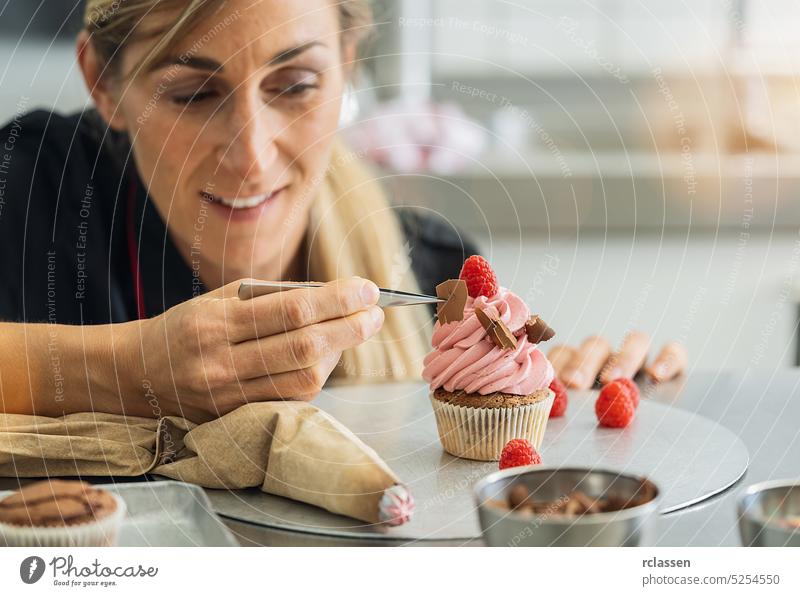 Glückliche Frauen in der Konditorei, als der Konditor mit einer Pinzette ein Stück Schokolade auf einem Muffin-Belag drapiert Spritzbeutel rosa Himbeeren Kuchen