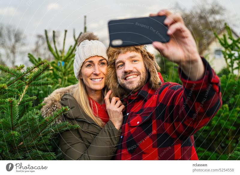 Frau und Mann halten Smartphone und lächeln, um ein Selfie auf dem Weihnachtsbaummarkt zu machen und Spaß zu haben Foto Telefon Fotokamera Kiefer Weihnachten