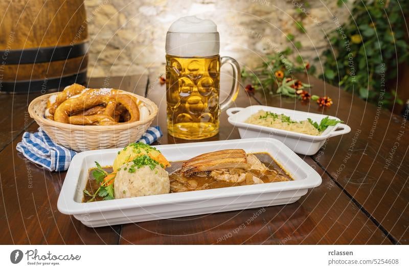 Schweinskrustenbraten mit Biersauce, in der deutschen Küche ein Schweinekrustenbraten mit Biersauce, mit Kraut- und Kartoffelknödel, Brezen und Bierkrug in einem bayerischen Biergarten oder auf einem Oktoberfest