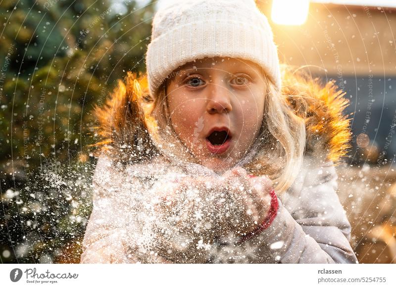 Kleines Mädchen Blowing Snow in frostigen Winter Park und lächelnd. Kind mit Spaß im Winter Zeit an Weihnachten jung Teenager Handschuhe Tanne Weihnachtsbaum