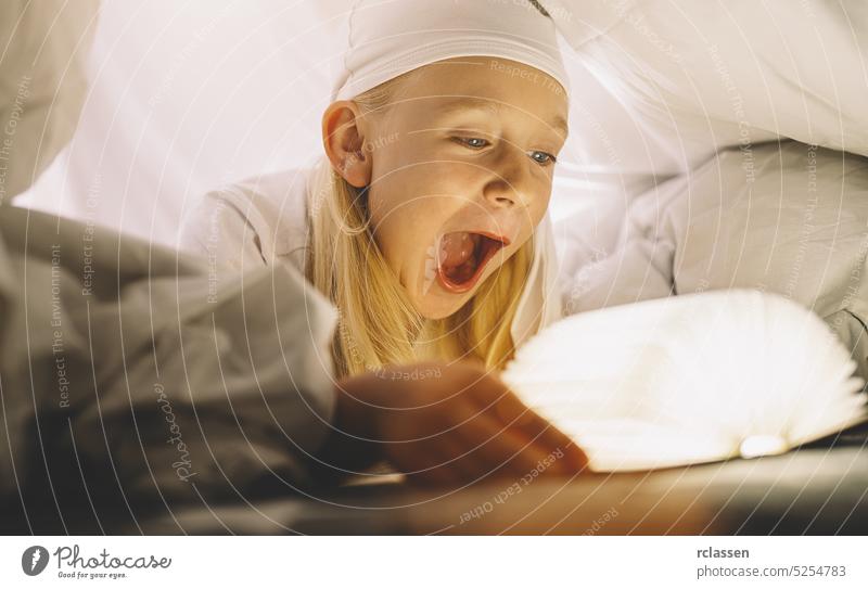 Erstauntes Kleinkind, das nachts unter einer Decke ein Buch liest. Magisches Licht, das aus dem Buch kommt. Kindheit Mädchen Taschenlampe Kinder Familie