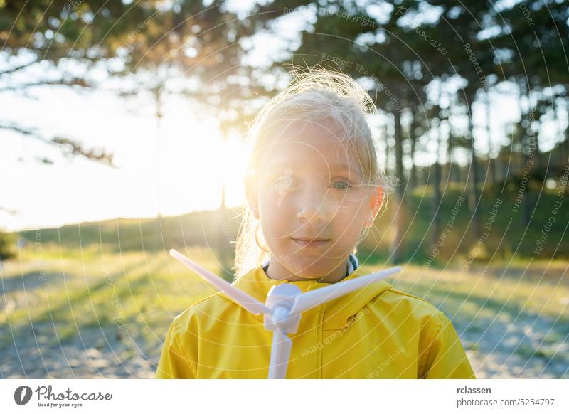 Blondes Kind hält Windradmodell bei Sonnenuntergang im Wald und schaut in die Kamera. Neue Energie Produktion Konzept Bild Alternative Energie Innovation