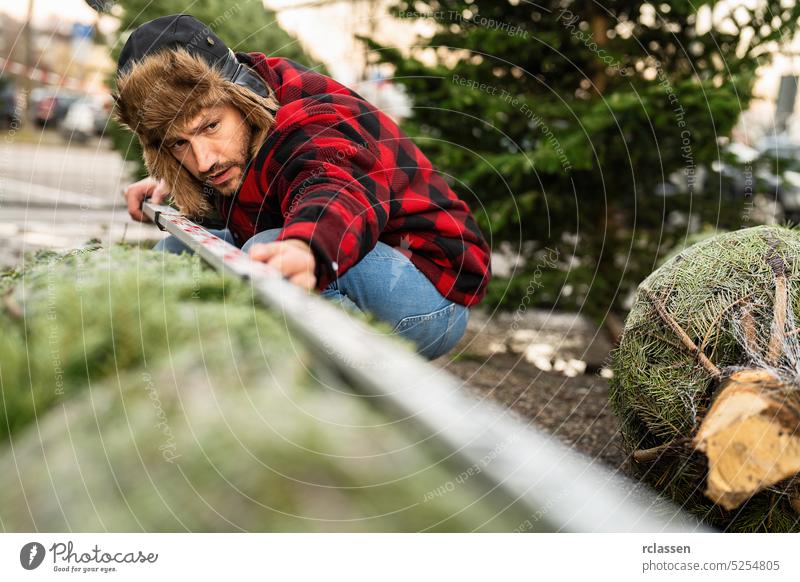 Mann, der auf einem Weihnachtsmarkt mit einem Lineal einen Weihnachtsbaum ausmessen und kaufen will prüfen Jeanshose Messgerät Kiefer Weihnachten Menschen Baum