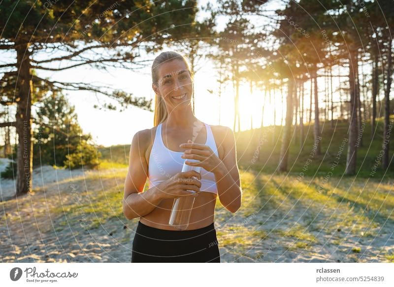 glückliche Frau Athleten nimmt eine Pause am Strand Wald, hält Wasserflasche und Blick auf die Kamera an einem heißen Tag. Sonnenaufgang trinken attraktive Frau