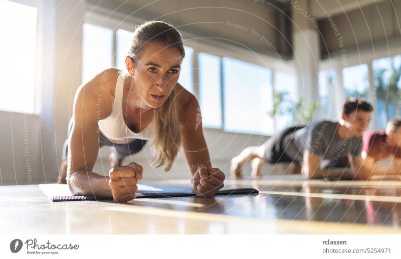 Fitness-Frau macht Planke Übung Workout in einer Gruppe von Menschen in der Turnhalle. Sport Mädchen und Männer in Sportkleidung tun Ausübung auf Yoga-Matte, Planken drinnen