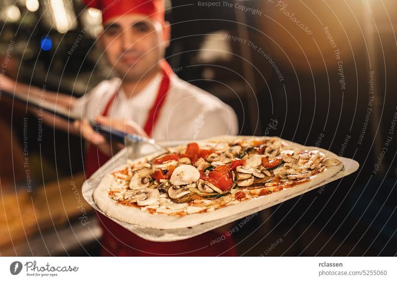 italienischer Pizzabäcker mit frischer Pizza auf der Schale in einer Pizzeria Teigwaren roh Holzfeuer Ziegelofen Chefkoch Feinschmecker professionell Arbeit