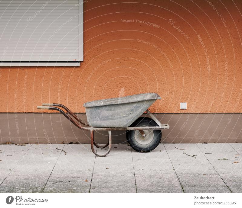 Schubkarre vor orangefarbener Wand mit geschlossener Jalousie als minimaoistisches Sinnbild für Hand- und Gartenarbeit Schiebkarre Karre Terrasse minimal