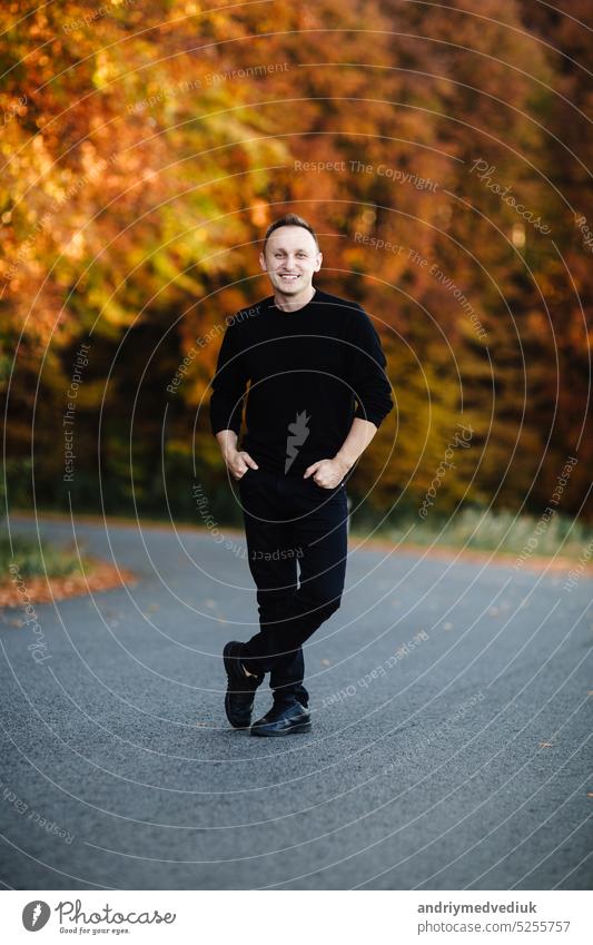 Junge attraktive Mann steht auf der Straße im Herbst Wald. Kerl mit kurzen Haaren trägt in schwarz ist in der Kamera im Park mit gelben Blättern Bäume suchen. Gemütlicher Tag auf dem Lande