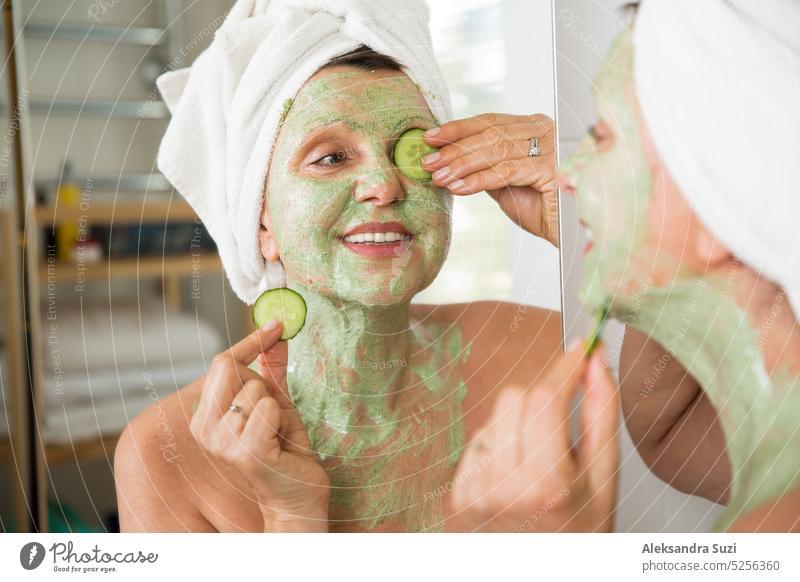 Reife schöne Frau Anwendung grün hausgemachte DIY Gesichtsmaske auf der Haut. Blick in den Spiegel im Bad, Spaß haben. Eingewickelt in ein Handtuch. Hautpflege-Routine und Anti-Aging-Behandlung für ältere Frauen.