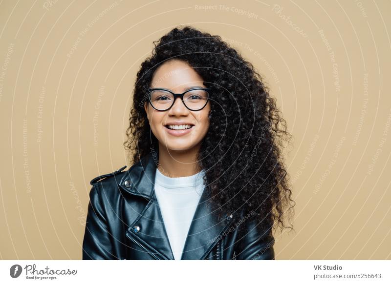 Stilvolle afroamerikanische Frau lächelt zähneknirschend, führt ein angenehmes Gespräch, genießt einen guten Tag, trägt eine durchsichtige Brille, Lederjacke, drückt positive Gefühle aus, posiert im Studio. Ethnizität, Mode
