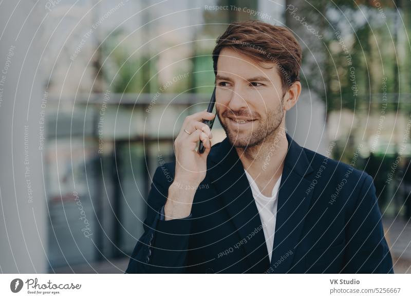 Gutaussehender, unrasierter, männlicher leitender Angestellter, der ein Geschäftsgebäude verlässt, spricht über sein Smartphone Business Mann